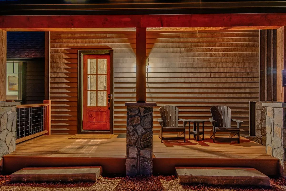 The image shows a welcoming red front door with wooden chairs on a large stone porch, warm porch lighting, and open space that hints at easy access to outdoor areas.