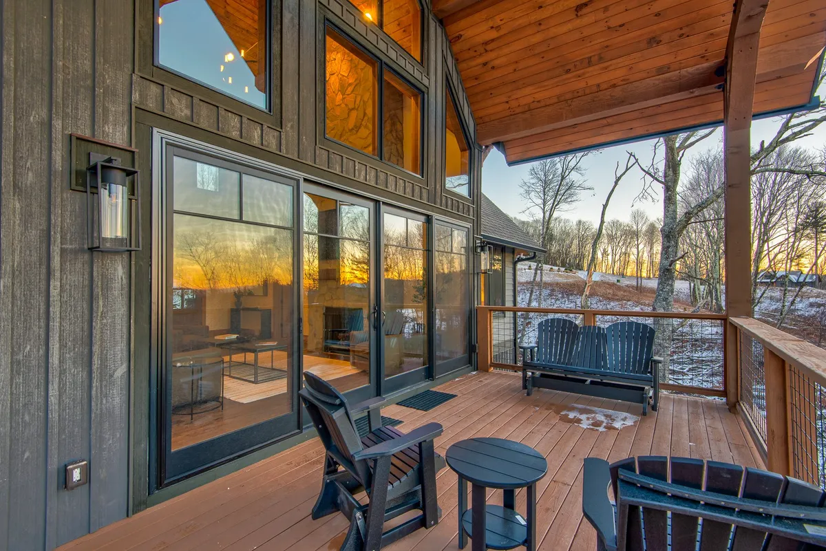 A spacious wooden deck with comfortable outdoor seating looks out over a beautiful sunset view. The large glass windows of the home glow with warmth, hinting at cozy gatherings both indoors and out. The fire pit and inviting seating create the perfect atmosphere for romantic evenings or small group get-togethers.