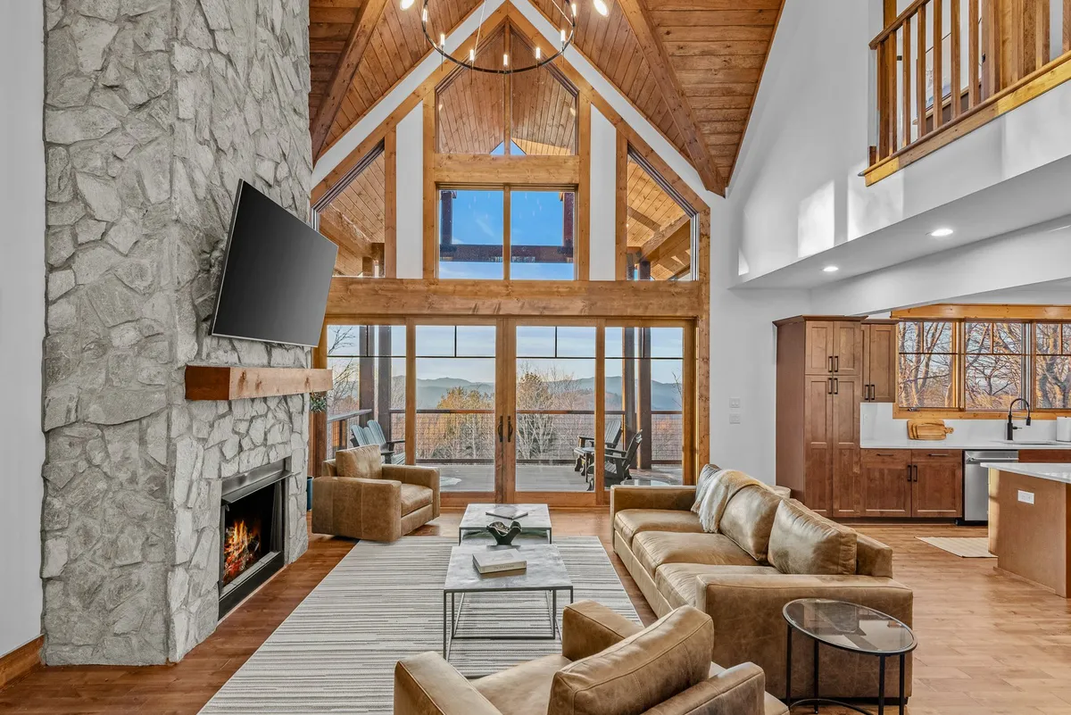A warm, inviting interior with a stone fireplace, large windows showcasing scenic views, a beamed ceiling, and comfortable seating. The space invites relaxation and social gatherings.