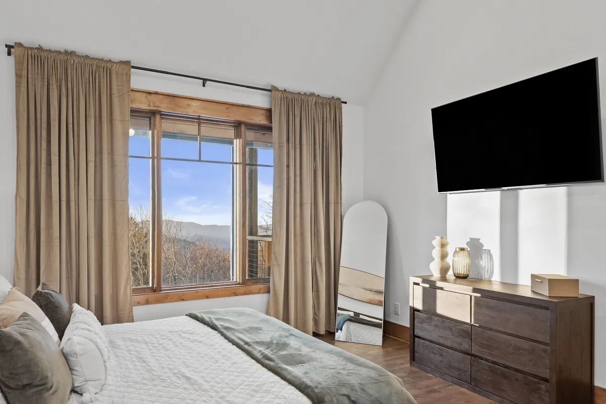 A cozy bedroom featuring a bed dressed in pillows and blankets, a large window framing a scenic mountain view, a wooden dresser, a large mirror, and a wall-mounted TV. The setup highlights opportunities for relaxation and entertainment.
