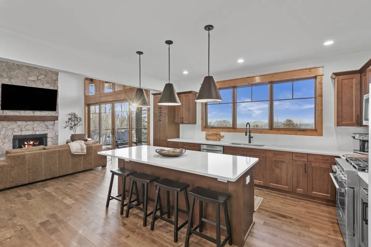 A wide-angle view of the open-concept living area, showcasing a plush leather sofa and swivel chairs gathered around a glowing gas log fireplace. The spacious granite kitchen island with barstools is visible in the background, connecting the living room to the kitchen seamlessly. Large windows bring in natural light, enhancing the inviting and celebratory atmosphere.