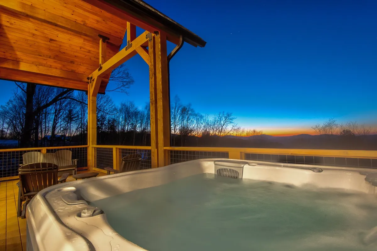 An inviting outdoor hot tub sits on a wooden deck, surrounded by tall trees with a stunning sunset sky in the background. The serene environment and natural setting create a peaceful retreat for couples or friends to relax and unwind together.