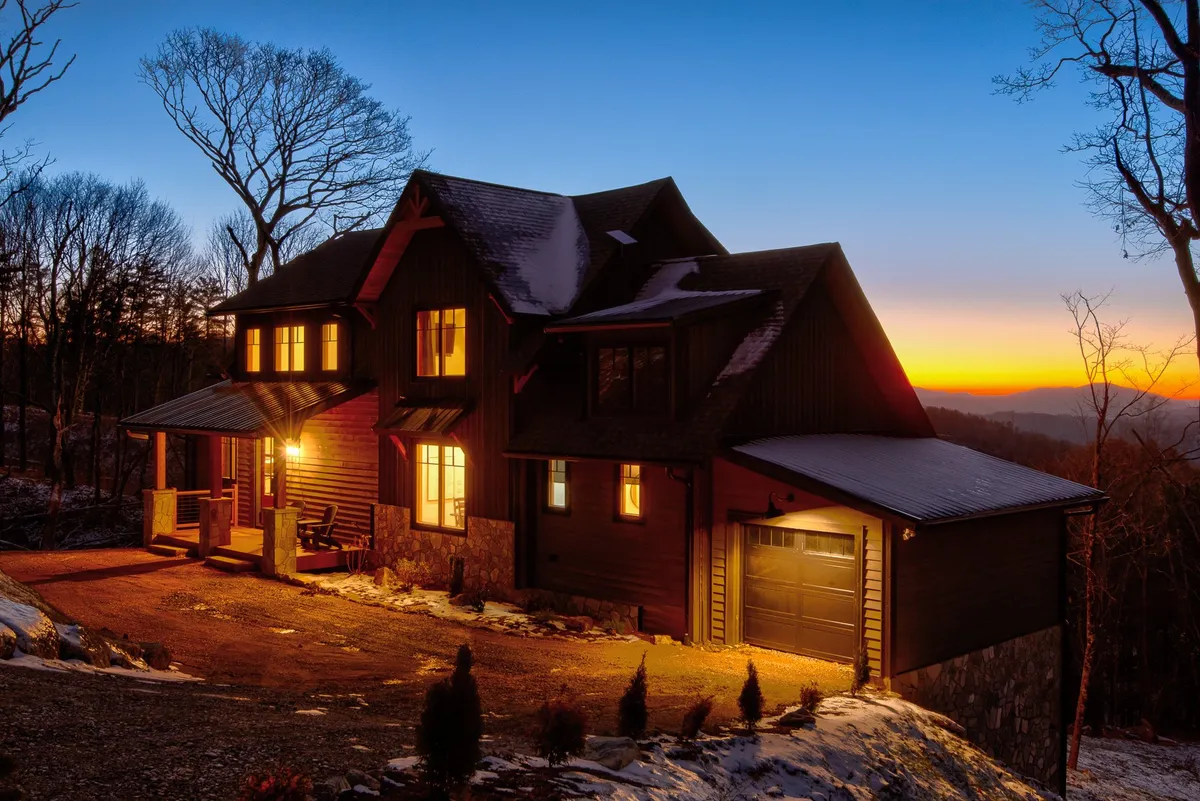 The home’s warmly lit windows and inviting porch seating are shown just at sunset, with nature trails and landscape visible in the background. The scene promises a cozy return after outdoor adventures and subtly suggests the presence of nearby hiking and gathering spaces.