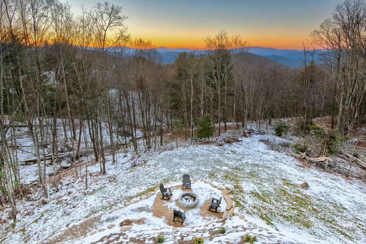An inviting outdoor area featuring a fire pit surrounded by Adirondack chairs, set against a backdrop of snow-covered ground, distant mountains, and tall trees. The scene exudes a cozy, communal atmosphere ideal for gatherings.
