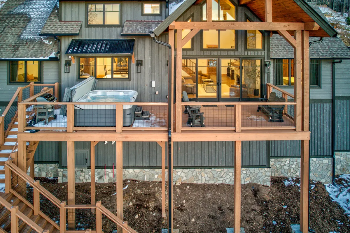 This image shows a wooden deck attached to the house, featuring a luxurious hot tub with ample seating and large windows overlooking the scenic mountain landscape. The deck is surrounded by trees and bathed in the warm light of sunset, creating a cozy and inviting atmosphere.