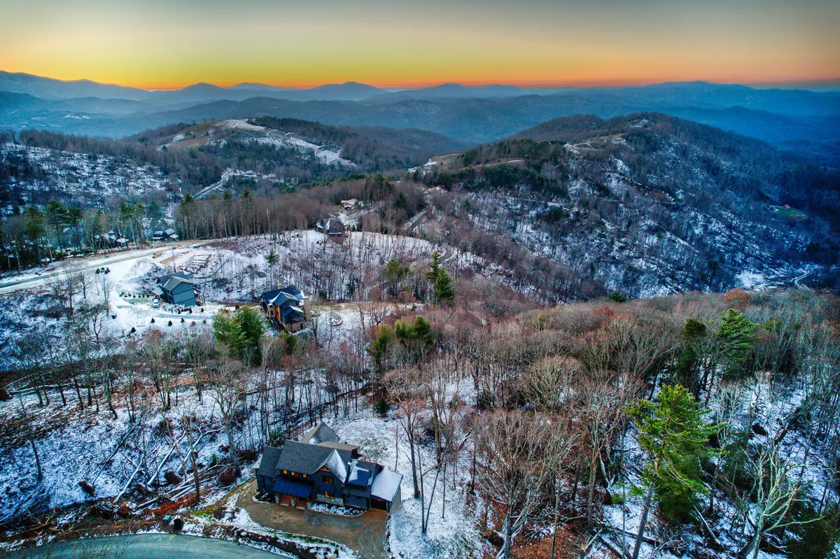 An expansive view of a mountainous landscape, blanketed in snow, with distant peaks and scattered homes visible. The scene evokes a sense of open space and opportunities for outdoor recreation, such as hiking, skiing, and exploring hidden trails. The sky hints at sunset, enhancing the adventurous appeal.