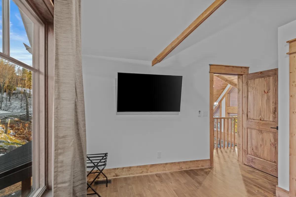 A comfortable lounge space with a window offering a view, a contemporary TV mounted on the wall, a wooden door, and cozy wooden floors. The arrangement suggests a flexible area for relaxing, watching TV, or serving as an extra sleeping space.