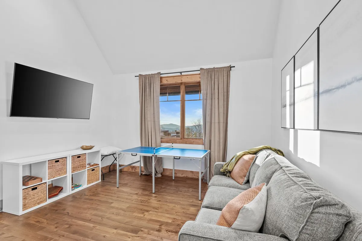 A playful interior space with a blue ping pong table, large window framing a mountain view, and a comfortable sofa. The area feels lively, perfect for fun and socializing.