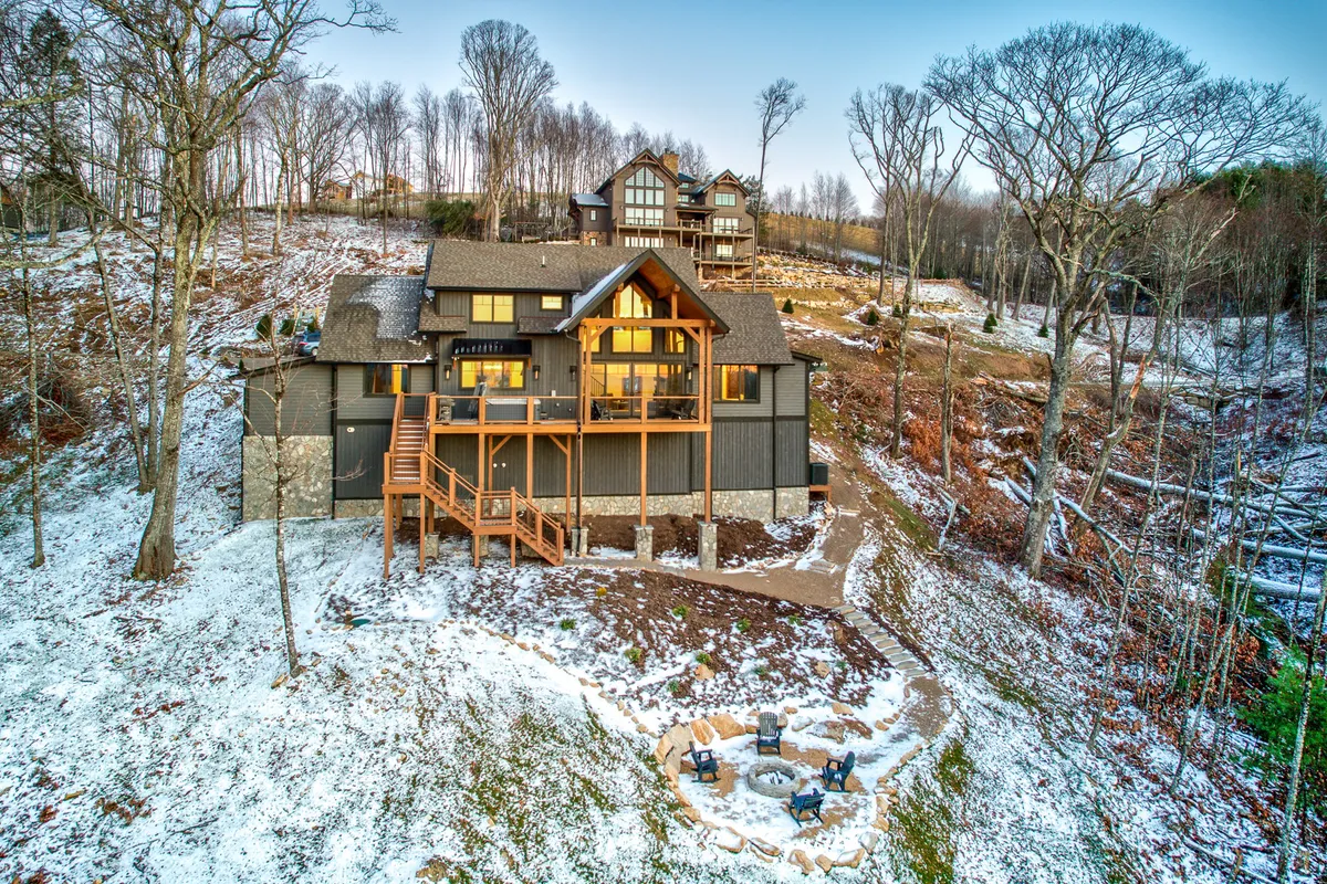 This perspective captures the large wooden deck extending from the home, with steps leading down to a private fire pit area surrounded by trees. The fire pit is set up for group gatherings with chairs around it, and the overall mood is cozy and inviting, perfect for evening relaxation and marshmallow toasting.