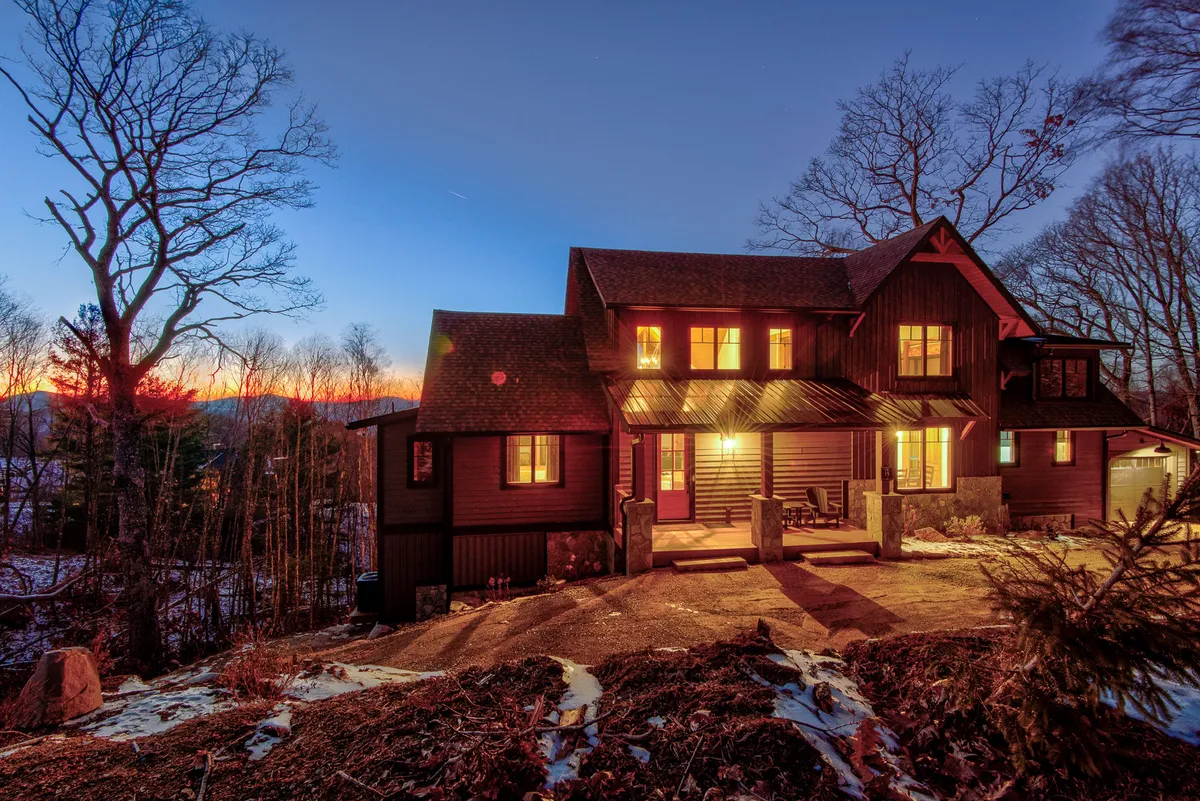 The image features the exterior of the home illuminated with warm lighting against a tranquil mountain sunset, surrounded by dark trees and open grounds. There's a visible fire pit area, suggesting a welcoming space to enjoy the outdoors with loved ones or pets.