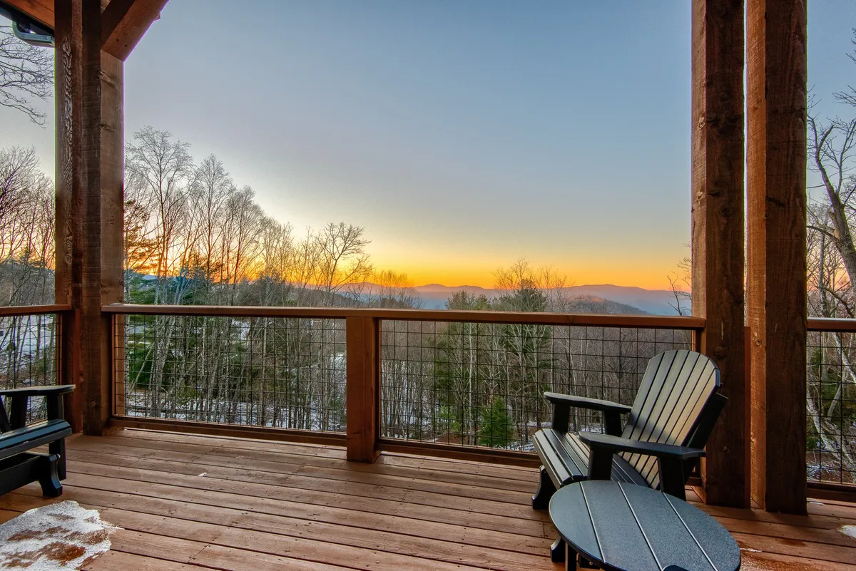 A tranquil scene of a wooden deck with Adirondack chairs facing a panoramic mountain view and a vibrant sunset sky. The deck looks ideal for sipping wine, enjoying conversation, or simply soaking in the serenity of nature.